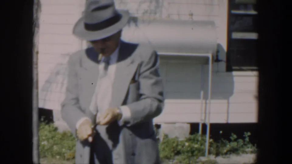 In Cuba 1951 footage, a suited man in a fedora stands outside a house, looking down while holding a cigar or adjusting his clothing.