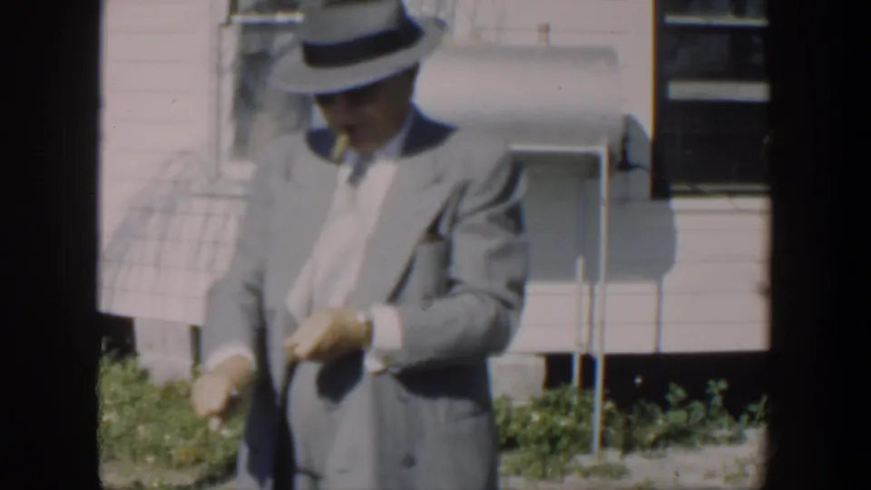 A man in a light suit and fedora appears in a 1951 Cuba home movie, standing outside a house while handling a cigar.