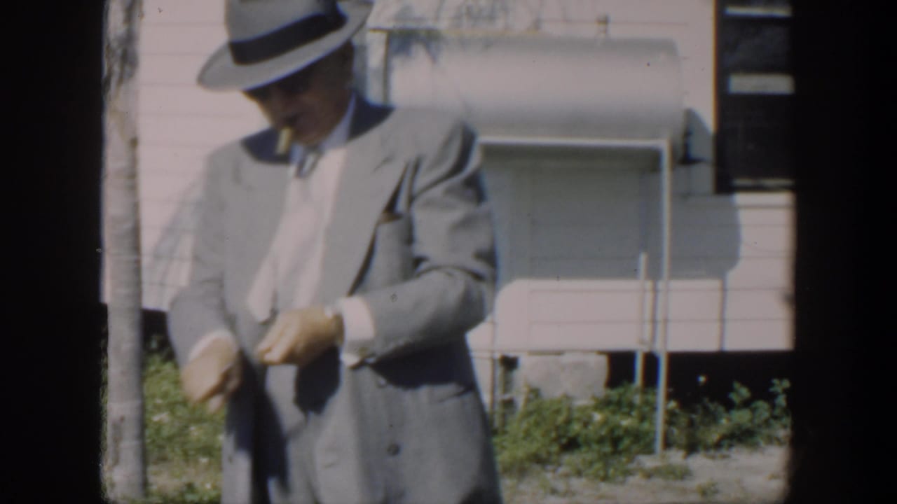 In Cuba 1951 home-movie footage, a man in a light gray suit and fedora stands outside a pale house, partly obscured by film-frame edges.