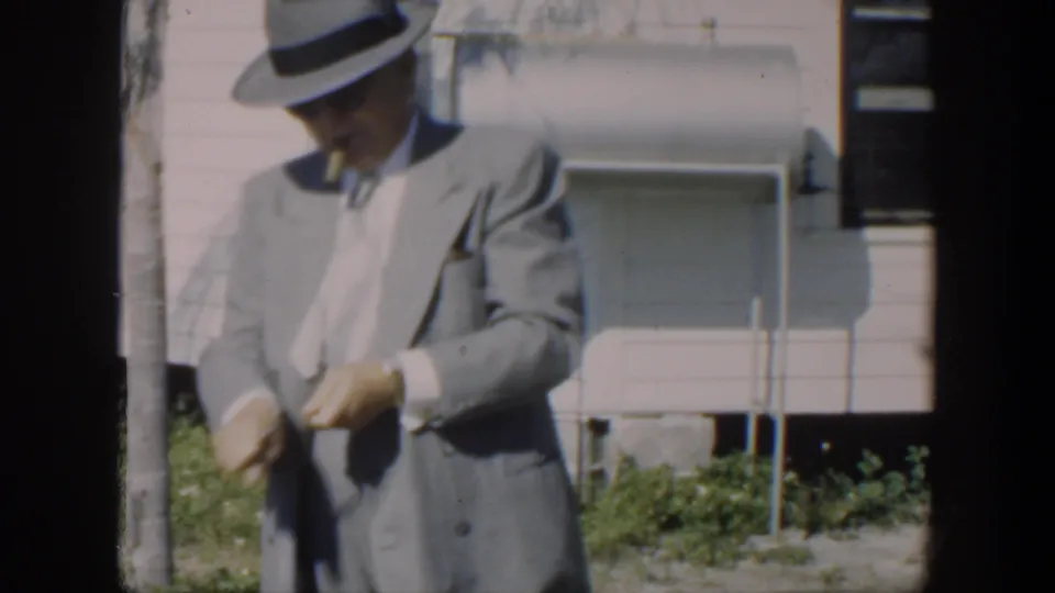 In Cuba 1951 home-movie footage, a man in a light gray suit and fedora stands outside a pale house, partly obscured by film-frame edges.