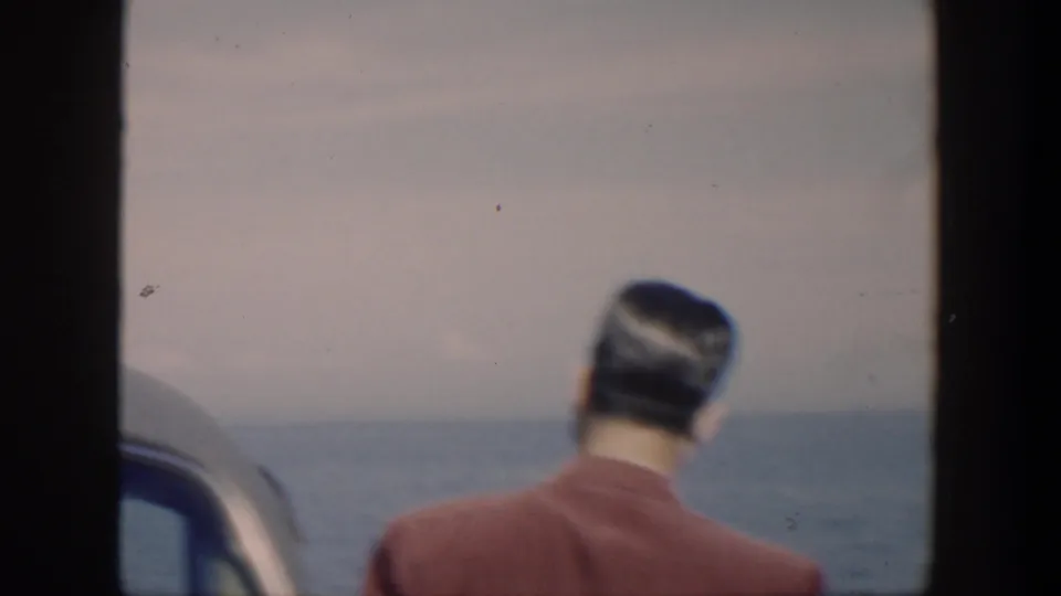 1951 Cuba home-movie frame of a man in a red shirt seen from behind near a car, looking toward the ocean.