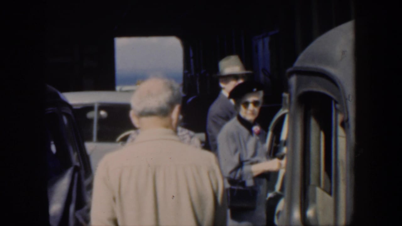Adults stand near classic cars in a shaded transport area.
