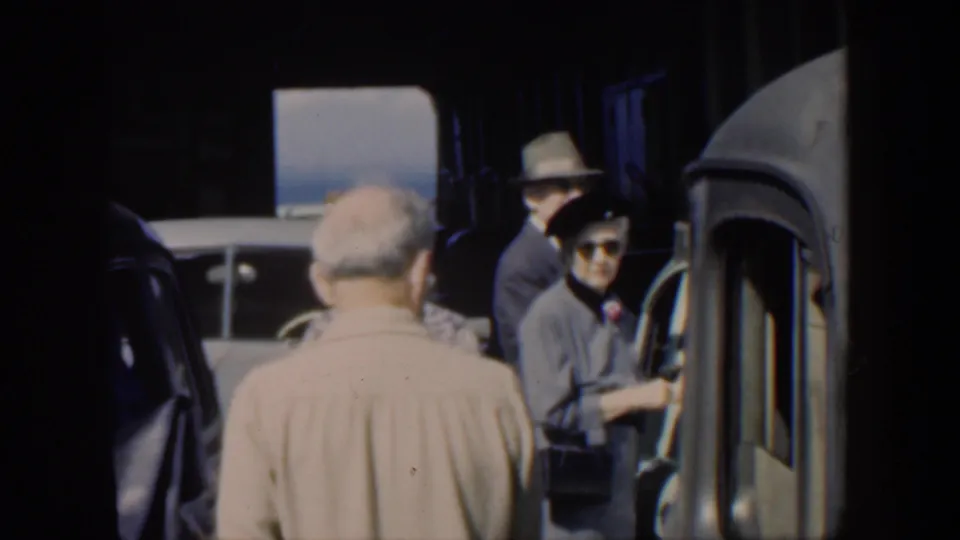 1951 Cuba home-movie frame showing adults in hats near rounded automobiles in a busy transport setting.