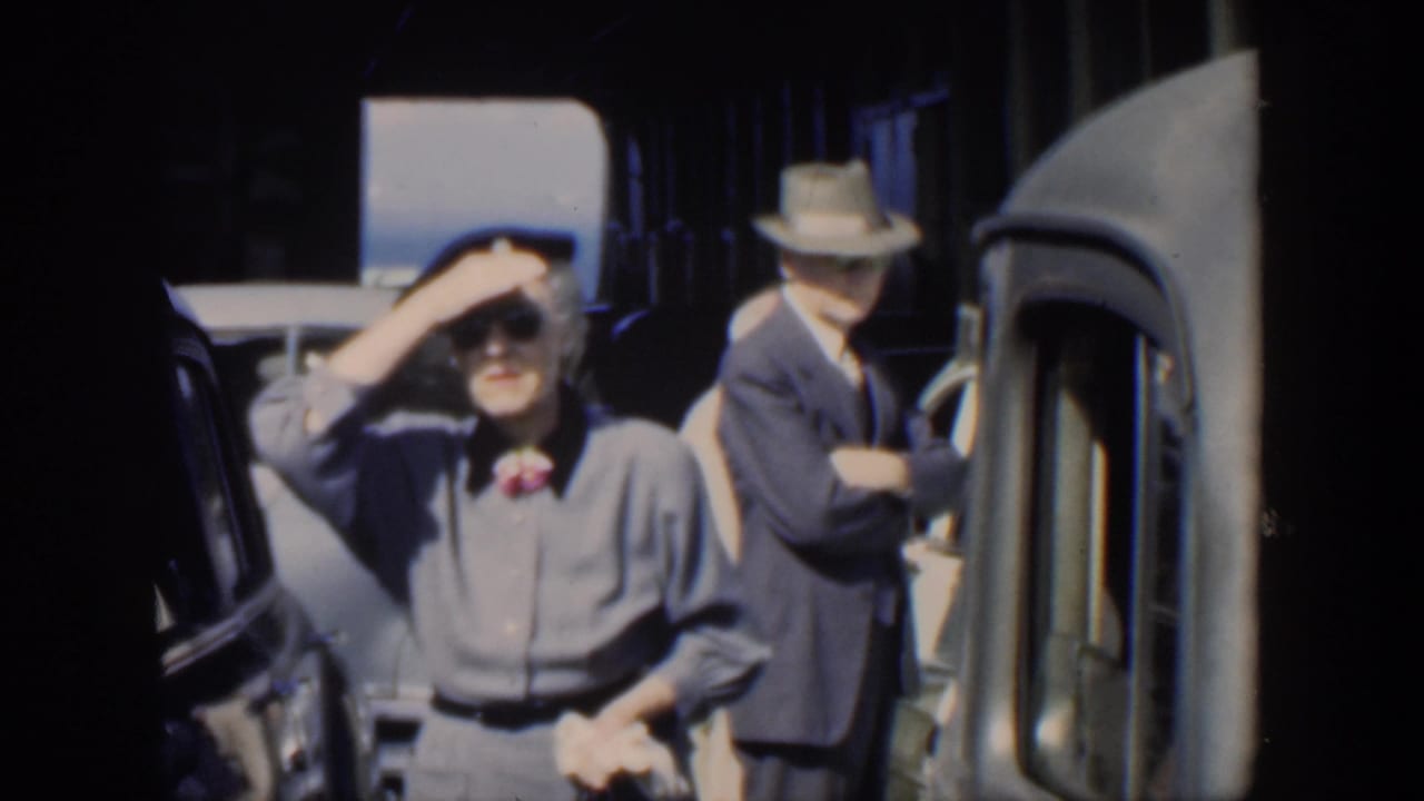 1951 Cuba home-movie frame showing a woman shielding her eyes beside classic cars while a suited man stands near a vehicle.