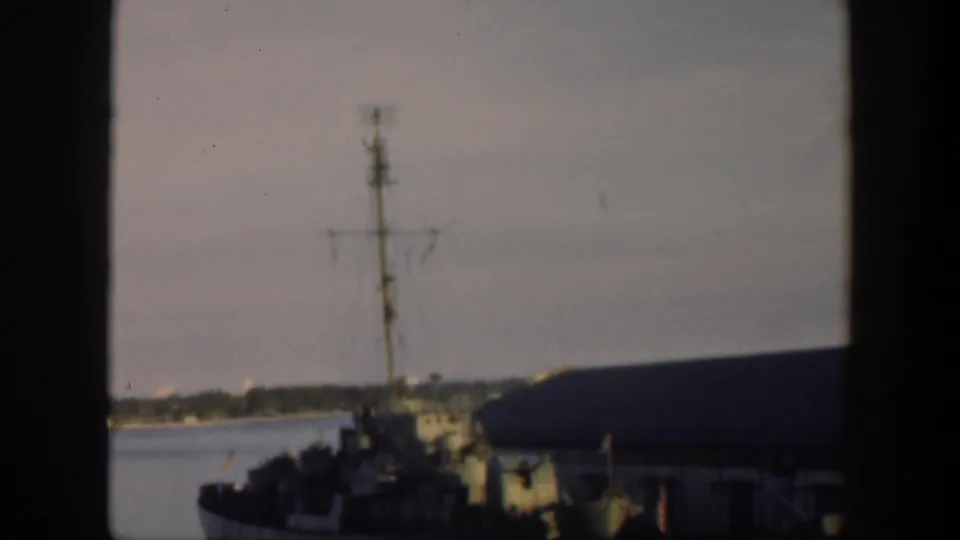 1951 Cuba frame showing the docked naval vessel beside a pier building, with mast rigging, harbor water, and overcast sky.