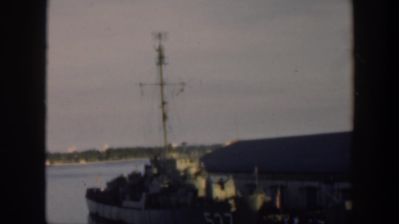 Distant gray naval ship partly framed by a dark window post at a harbor pier.