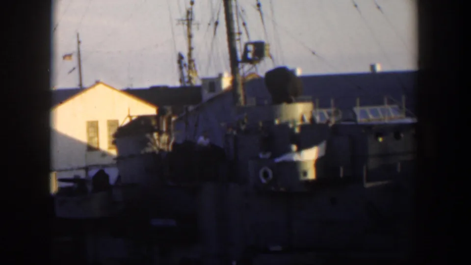 1951 Cuba frame showing the rear section of a docked naval vessel, rigging, a small flag, and waterfront buildings in low sun.