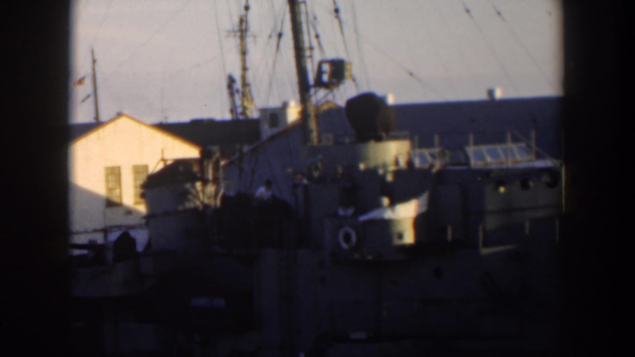 Ship superstructure and rigging at an industrial waterfront with a pale building behind it.