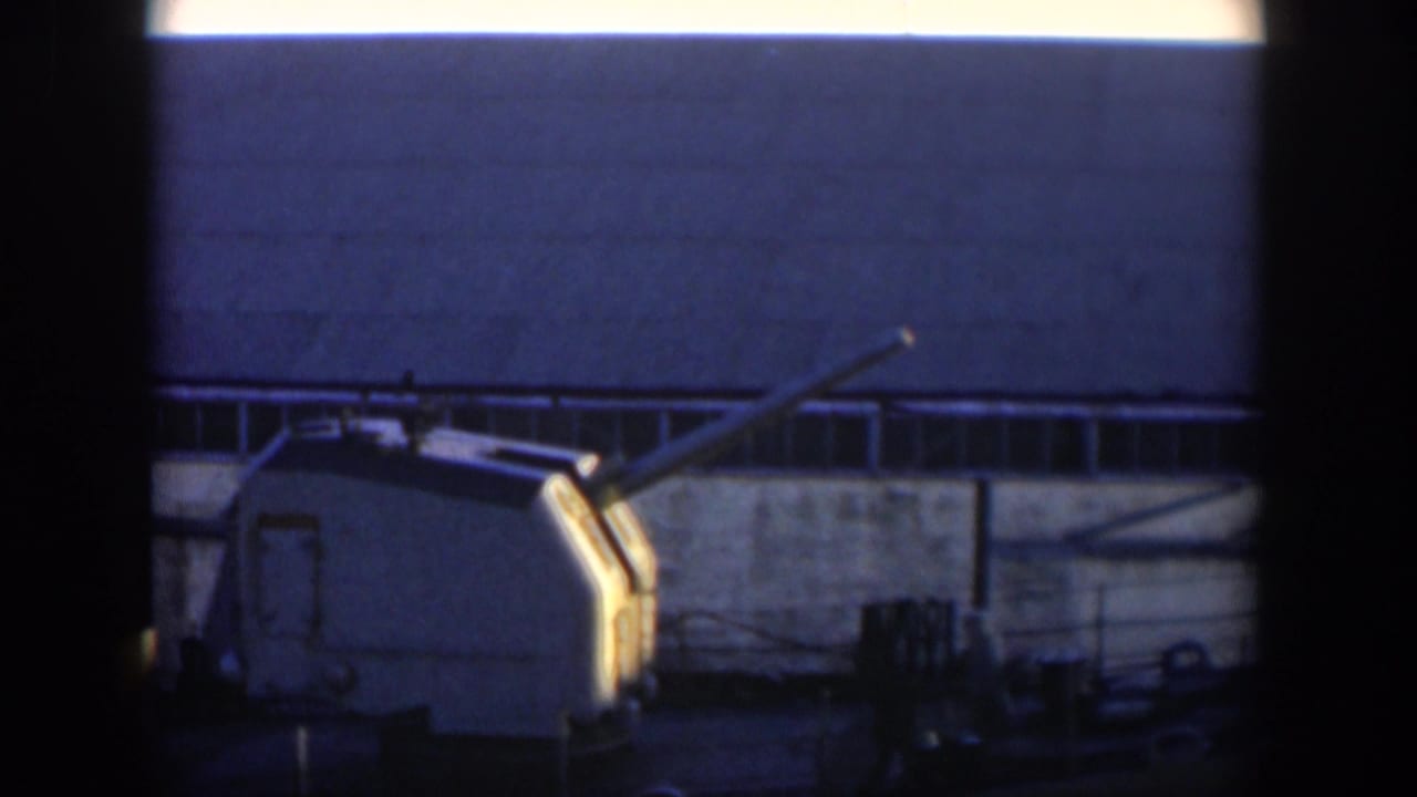 Naval gun turret on a ship deck beside an industrial building, partly framed by dark film edges.