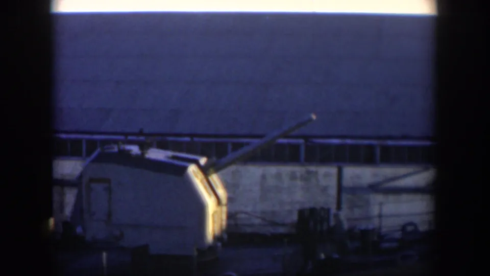 1951 Cuba 8mm frame showing a shipboard naval gun turret, deck railings, cables, and an industrial building behind it.