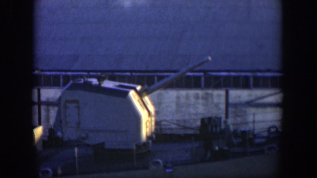 Shipboard gun turret with long barrel angled upward against an industrial building wall.