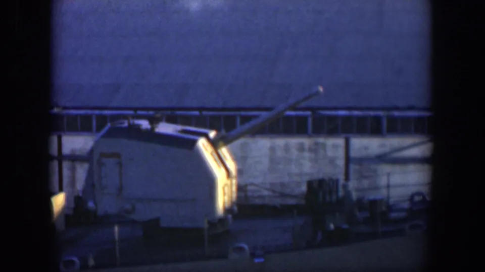 1951 Cuba 8mm frame with a naval turret on deck, sunlight catching the turret edge, and a large industrial building behind.