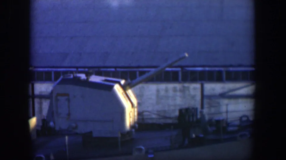 1951 Cuba 8mm frame of a ship deck turret and angled barrel, with industrial walls, cables, and railings in the background.