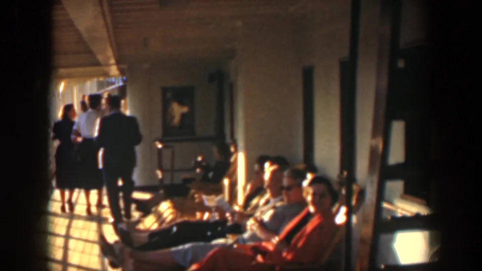 1951 Cuba frame showing a hospital or clinic corridor with seated adults in foreground and visitors walking away in warm light.