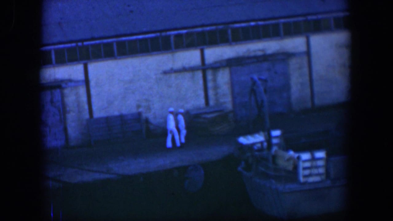 Two sailors walk on a dock beside a moored boat and warehouse wall.