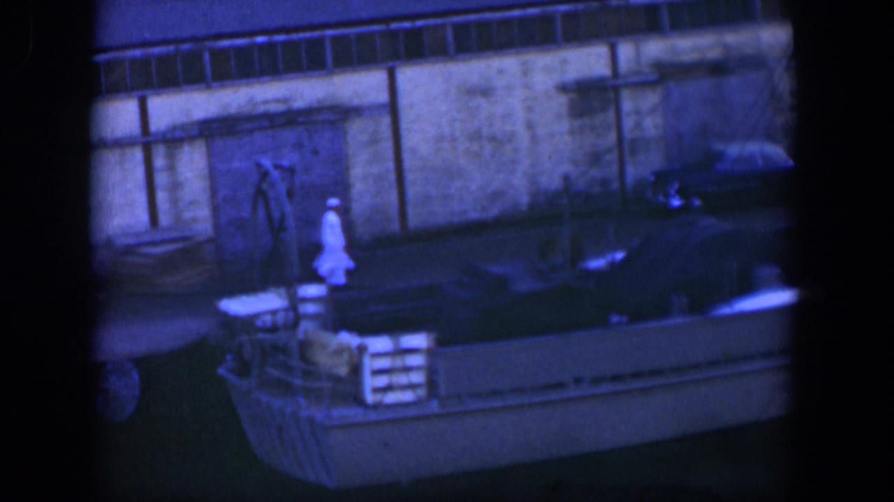 Man in a white uniform walks on a dock beside a moored boat and wall.