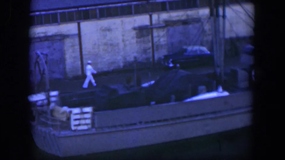 A man in white continues walking on a Cuban dock in 1951, with a moored boat, wall and dark parked car visible nearby.