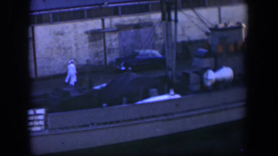The dockside scene shows the man in white near barrels and a moored boat, with a parked car and building wall in the background.
