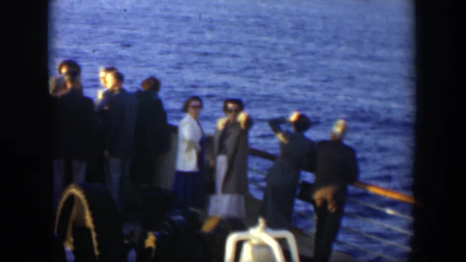 Adult passengers aboard a ship, some wearing sunglasses and coats, stand near the rail with blue ocean visible beyond them.