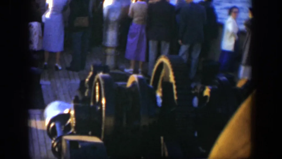 The frame emphasizes ship deck equipment in the foreground while adult passengers stand near the rail against the ocean backdrop.
