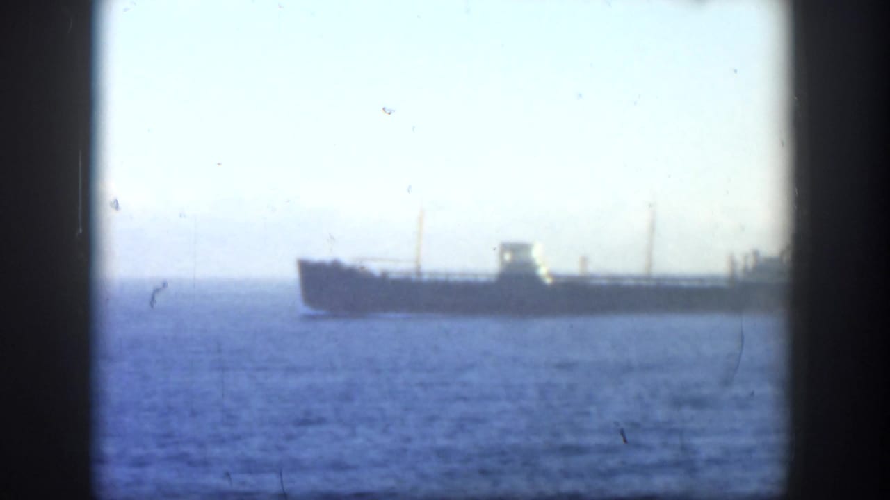 Distant cargo ship crossing calm blue water under a pale sky, seen through dark frame edges.