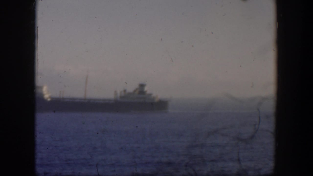 Distant ship crossing blue-gray water under a hazy sky, seen through a dark window frame.