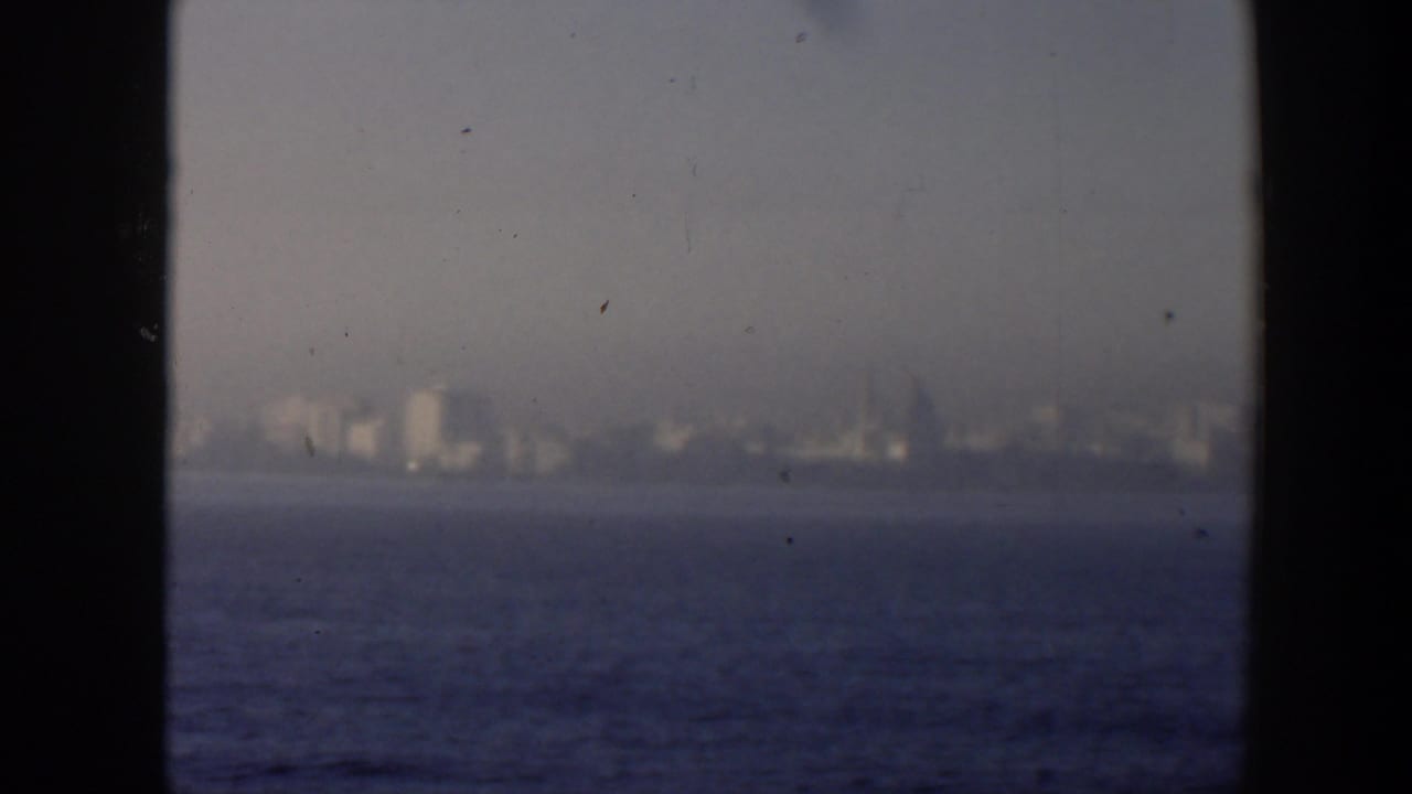 1951 Cuba 8mm frame showing a soft distant waterfront skyline across blue ocean, seen through dark window or rail edges.