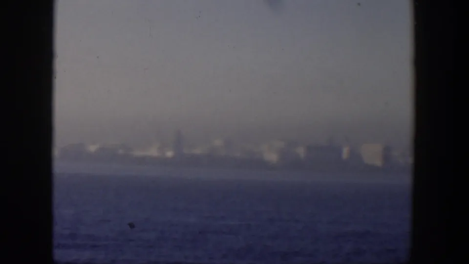 Soft 1951 Cuba coastal view with distant pale buildings, blue ocean foreground, haze, and dark film-frame edges.