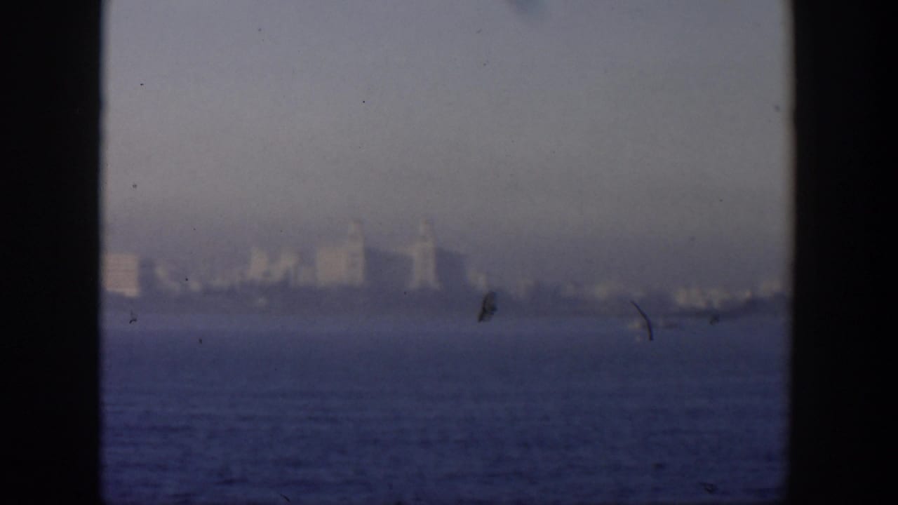 Hazy distant city skyline across blue water, framed by dark film borders.