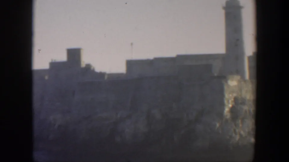 1951 Cuba 8mm frame showing a hazy coastal fortification with a tall lighthouse or tower rising near the right edge.