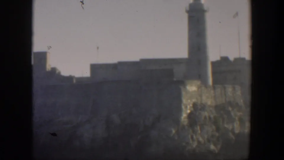1951 Cuba frame with a tall lighthouse or tower beside fortress walls, viewed through dark foreground framing and haze.