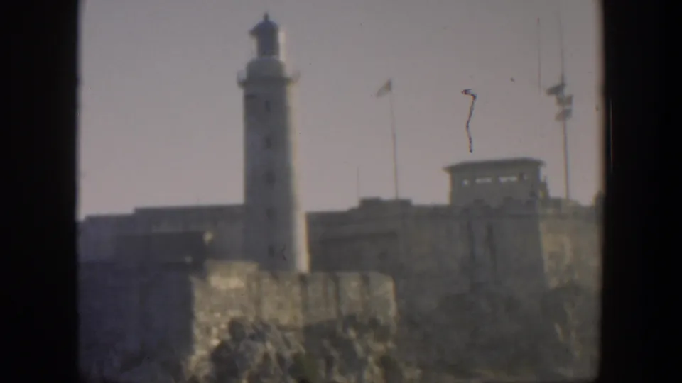 1951 Cuba 8mm view of a tall lighthouse, adjacent fortress buildings, flagpoles, and rugged stone foreground.