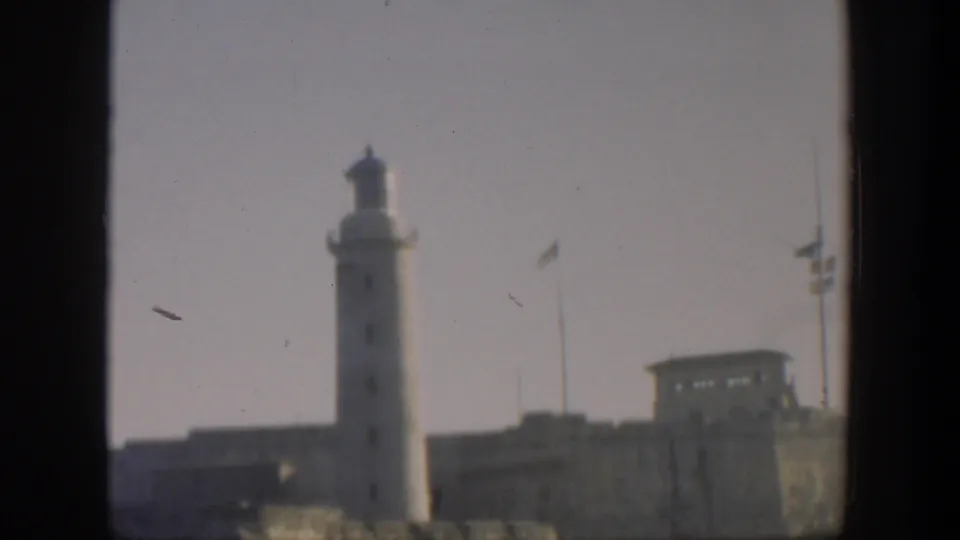 1951 Cuba frame showing the lighthouse, fortified structures, flagpoles, and pale sky, with a dark film edge across the bottom.