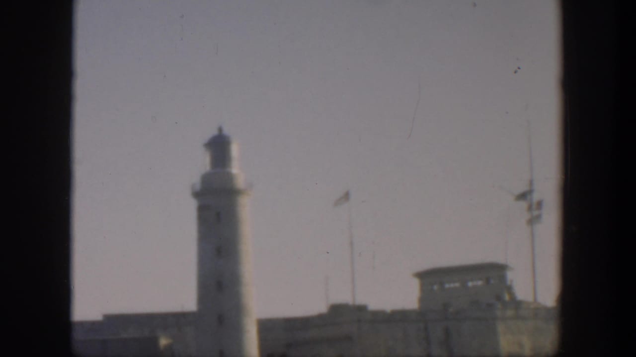 1951 Cuba 8mm frame showing a white lighthouse above a fortified wall, with flagpoles and dark contact-sheet borders.