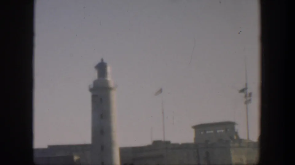 1951 Cuba 8mm frame showing a white lighthouse above a fortified wall, with flagpoles and dark contact-sheet borders.