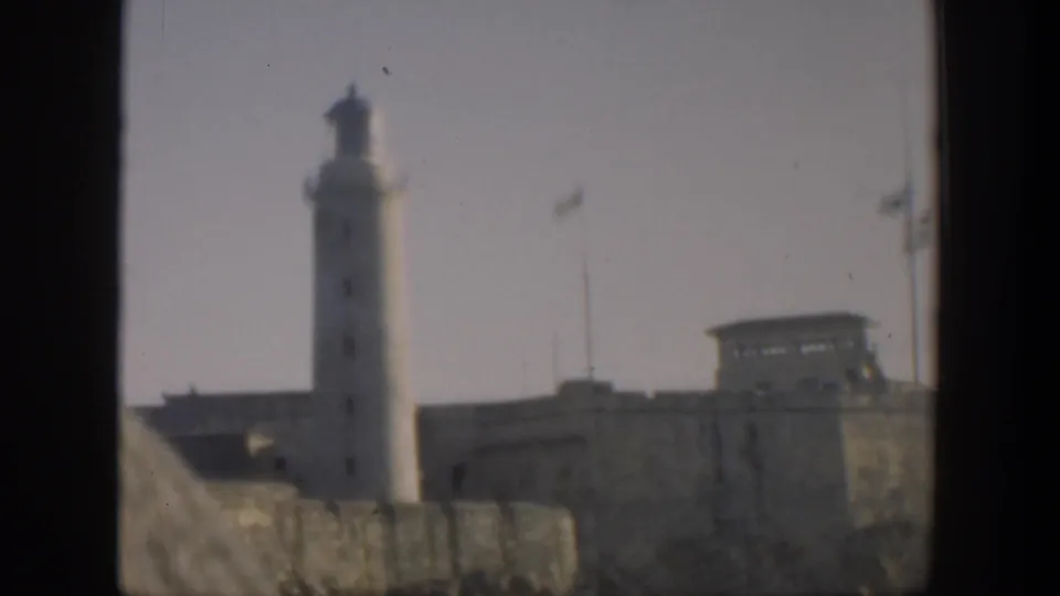 1951 Cuba home-movie frame with a tall lighthouse, masonry walls, roof structure, flagpoles, and soft faded color.