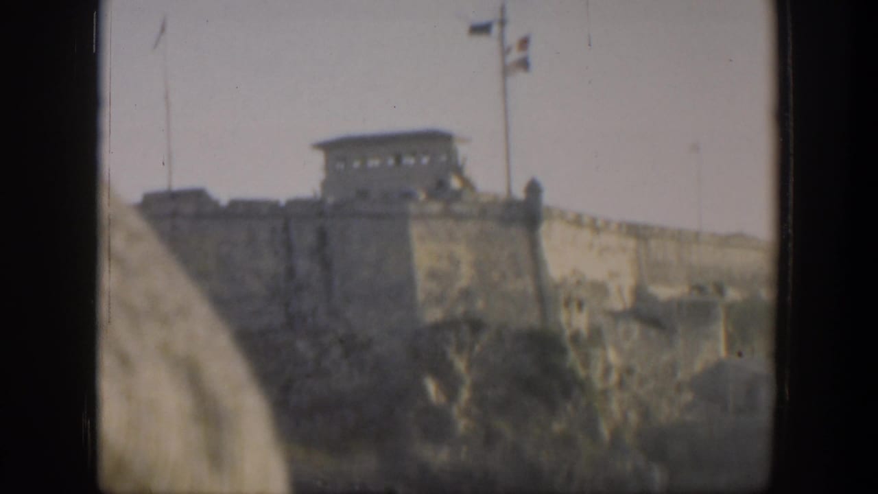1951 Cuba home-movie frame showing a stone fortress on high ground above the water, with flags and heavy gate-edge shadows.