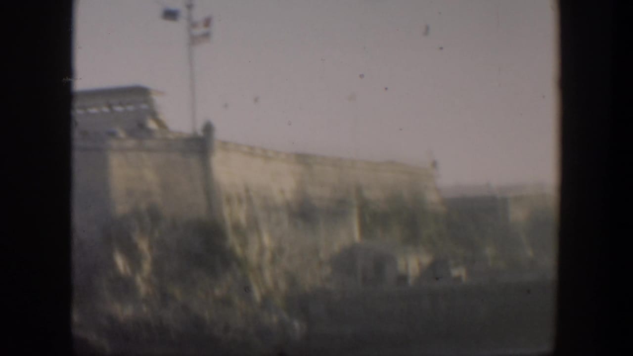 Fortress wall and tower on high ground, with flagpoles visible against a hazy sky.