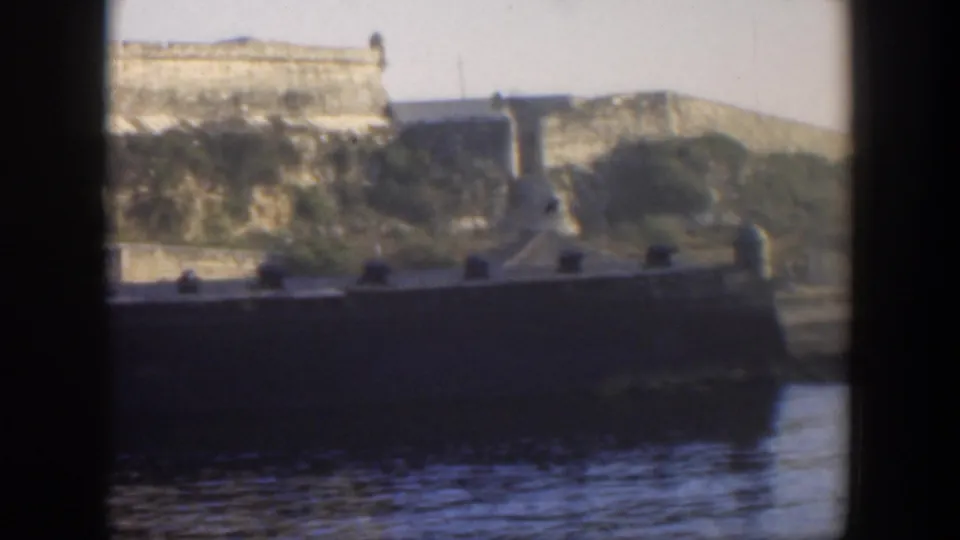 1951 Cuba frame with water in the foreground, a dark shoreline wall or dock, and sunlit fortress masonry on the rocky high ground.
