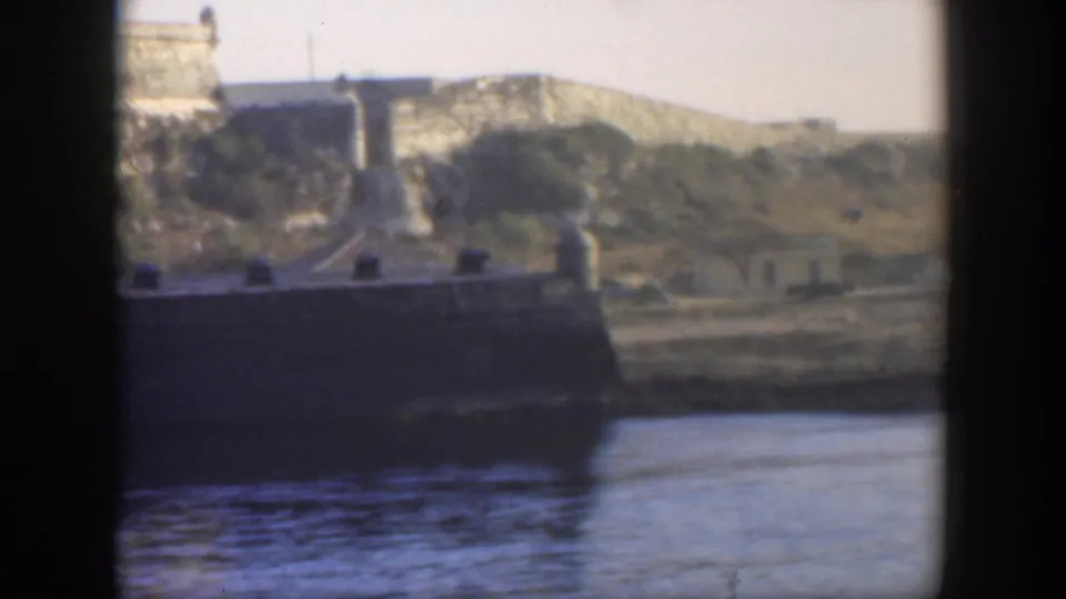 Late frame from 1951 Cuba footage showing water, a dark waterfront structure, and fortress walls stretching across the high ground.