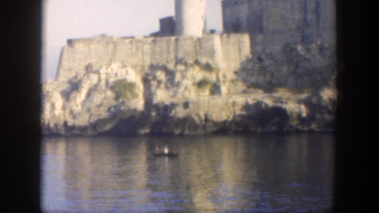 1951 Cuba 8mm frame showing a small boat on calm water near rocky shoreline, tan fortress walls, and a white lighthouse.