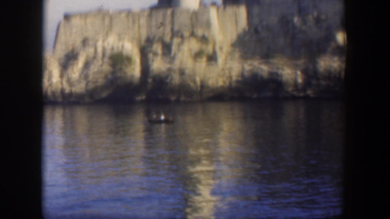 Fortress wall and rocky shore reflected in calm water with a small boat offshore.