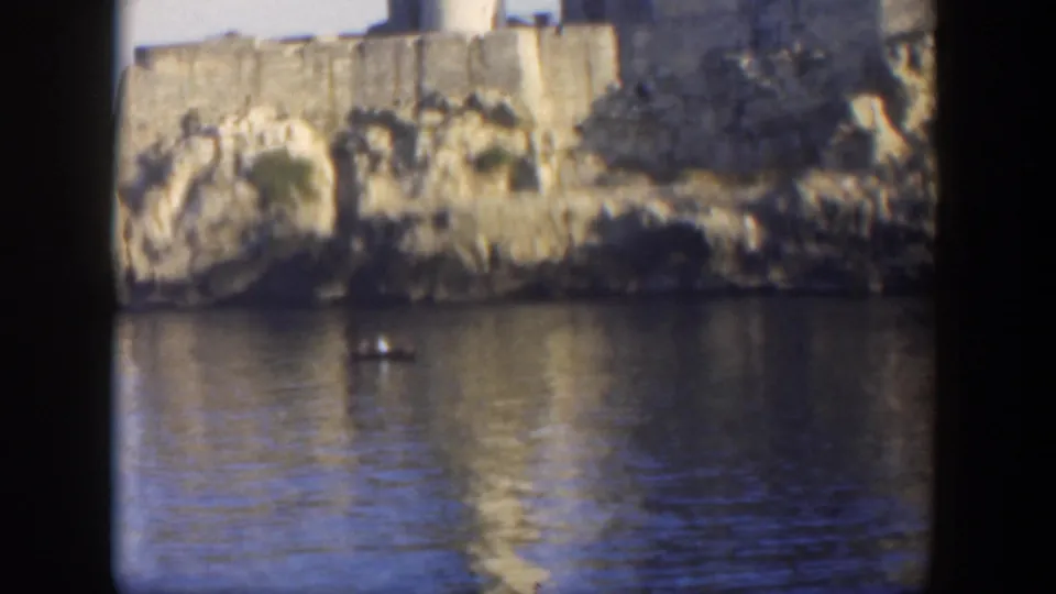 1951 Cuba home-movie frame of a small boat moving across calm water below weathered tan fort walls and shoreline rocks.