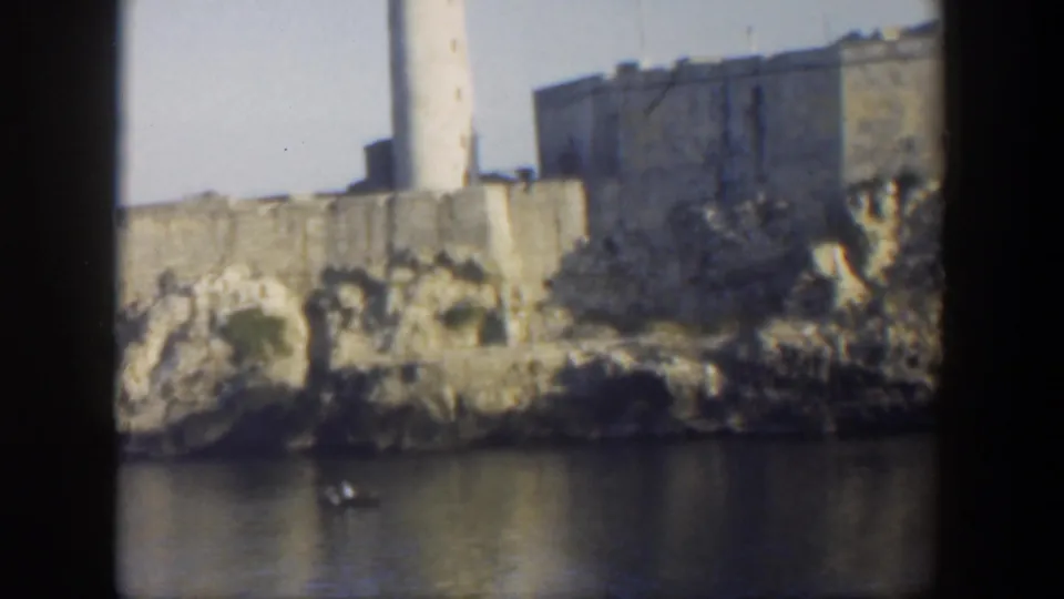 1951 Cuba 8mm frame showing a white lighthouse rising above tan fortress walls with a small boat in the water below.
