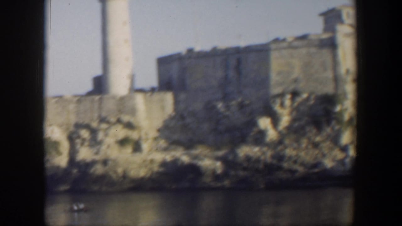 1951 Cuba home-movie frame showing a stone coastal fortress, lighthouse, rocky edge, and water in soft 8mm focus.