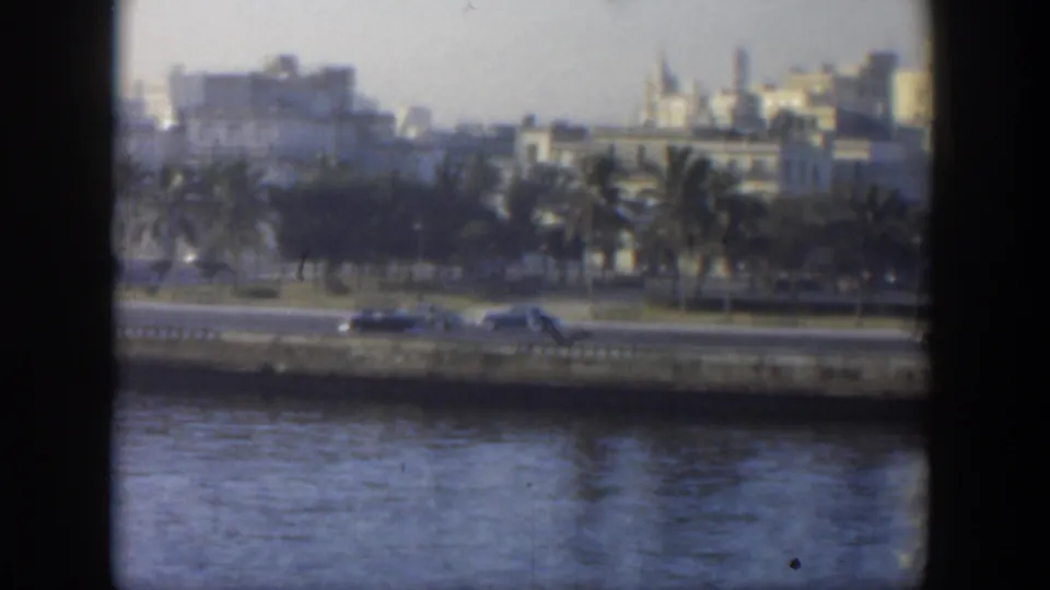 1951 Cuba home-movie frame showing cars along a waterfront road, palms, water, and a distant urban skyline.