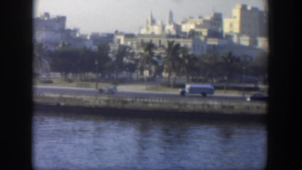 1951 Cuba waterfront traffic scene with a white bus and cars moving past palms and pale city buildings.