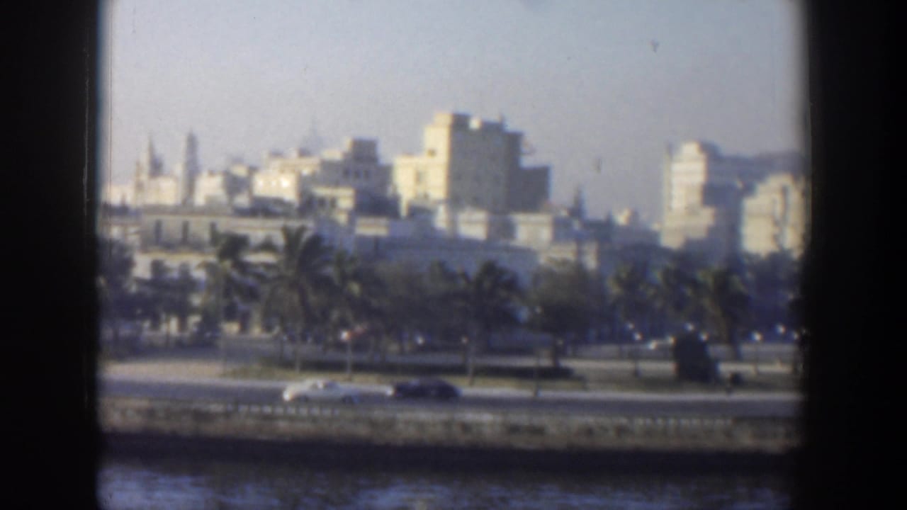 1951 Cuba waterfront view through a dark frame, with cars on a coastal road, palms, and pale urban buildings beyond.