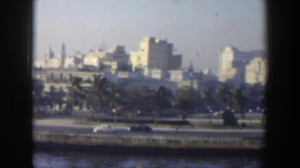 1951 Cuba waterfront view through a dark frame, with cars on a coastal road, palms, and pale urban buildings beyond.