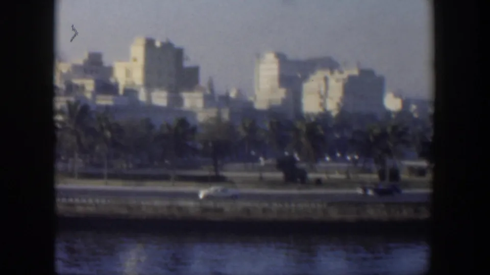 1951 Cuba city waterfront seen through a window-like divider, with palms, moving cars, and pale mid-century buildings.